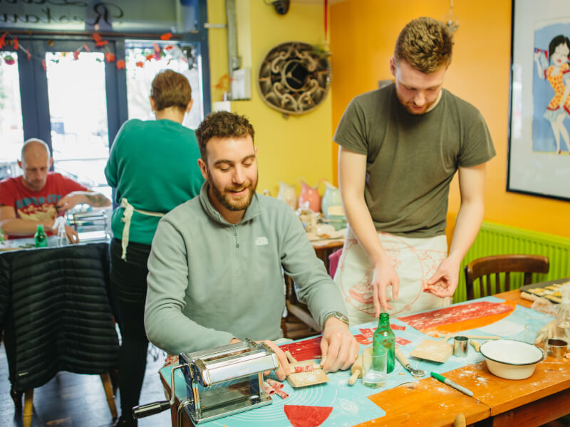 Two men rolling pasta, one sitting and one standing.